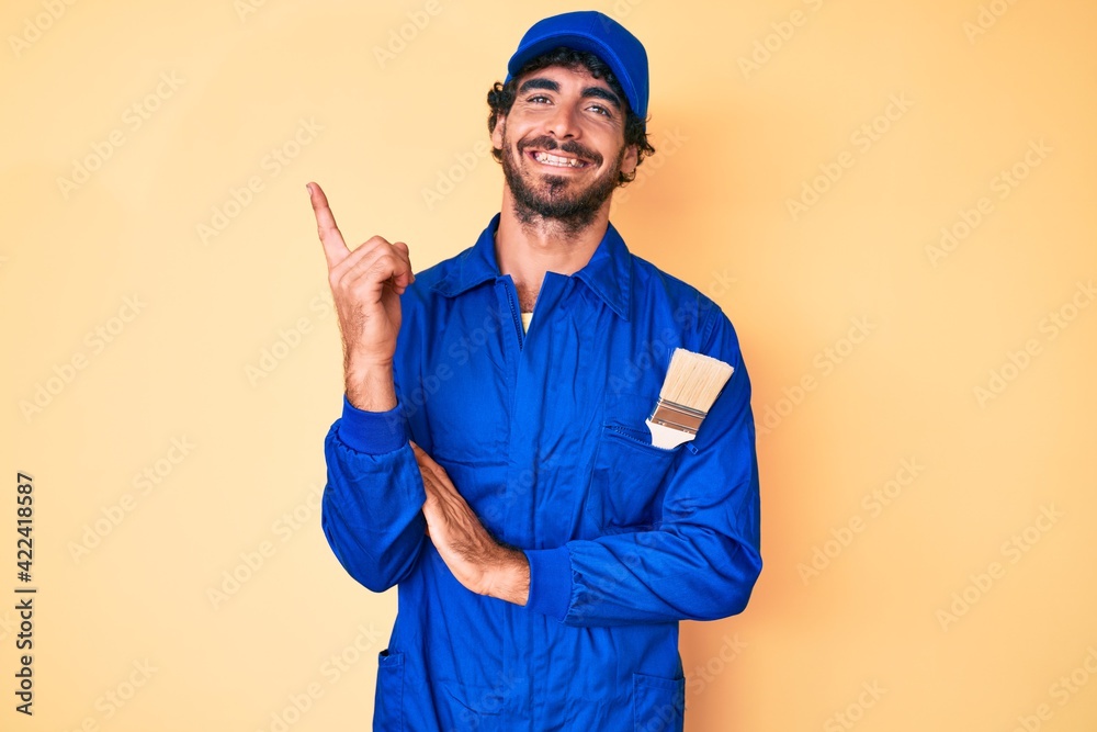 Handsome young man with curly hair and bear wearing builder jumpsuit uniform with a big smile on face, pointing with hand and finger to the side looking at the camera.