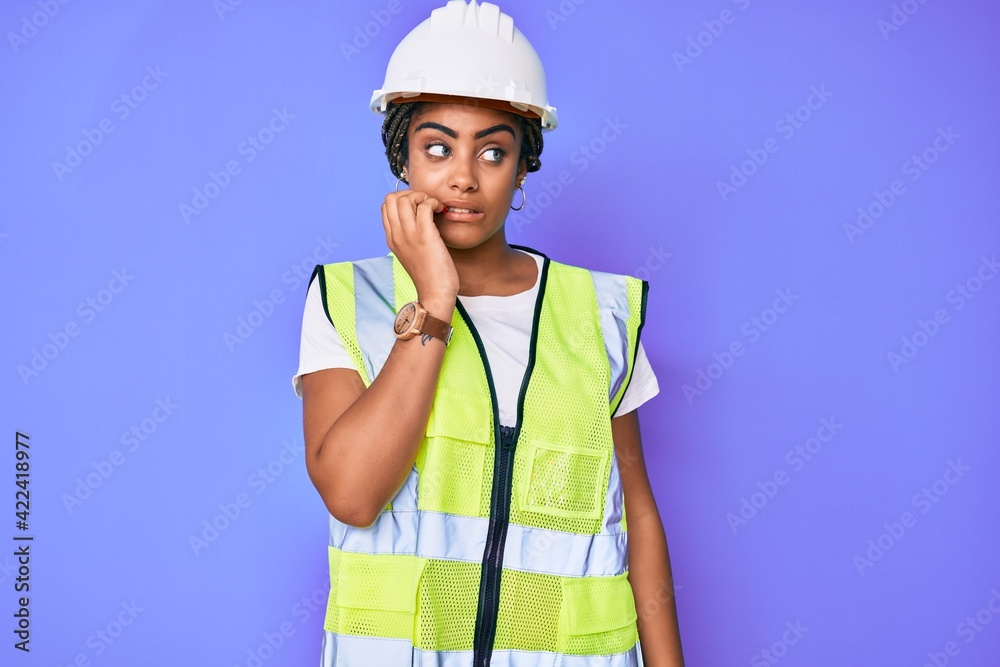 Young african american woman with braids wearing safety helmet and reflective jacket looking stressed and nervous with hands on mouth biting nails. anxiety problem. Young african american woman with braids wearing safety helmet and reflective jacket looking stressed and nervous with hands on mouth biting nails. anxiety problem.
