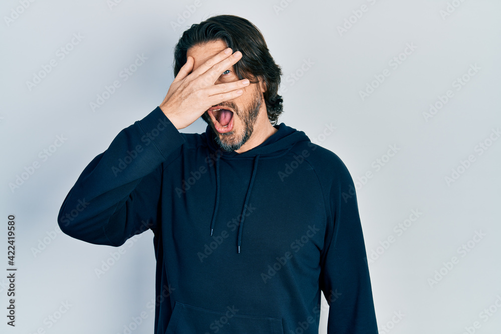 Middle age caucasian man wearing casual sweatshirt peeking in shock covering face and eyes with hand, looking through fingers with embarrassed expression. Middle age caucasian man wearing casual sweatshirt peeking in shock covering face and eyes with hand, looking through fingers with embarrassed expression.