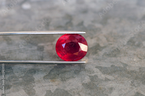 Deep saturated red natural mined Madagascar ruby, corundum - Al2O3 gemstone. Heated, treated, lead glass filled. Holded in tweezers. Clarity - Si,I2. Background - gray metal zinc pattern. For jewelry.