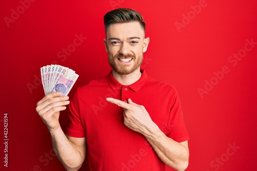 Young redhead man holding 20 polish zloty banknotes smiling happy pointing with hand and finger