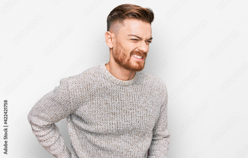 Young redhead man wearing casual winter sweater suffering of backache, touching back with hand, muscular pain Young redhead man wearing casual winter sweater suffering of backache, touching back with hand, muscular pain