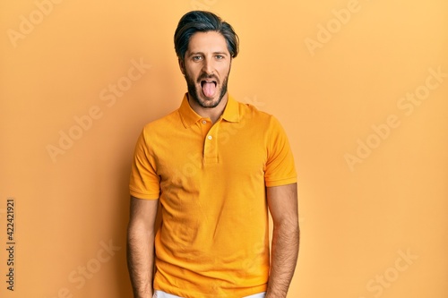 Young hispanic man wearing casual yellow t shirt sticking tongue out happy with funny expression. emotion concept.