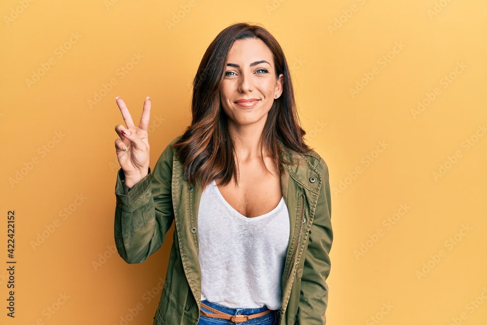 Young brunette woman wearing casual clothes over yellow background smiling with happy face winking at the camera doing victory sign. number two.