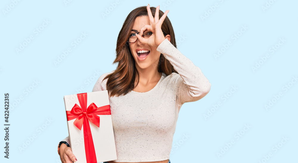 Young brunette woman holding gift smiling happy doing ok sign with hand on eye looking through fingers Young brunette woman holding gift smiling happy doing ok sign with hand on eye looking through fingers