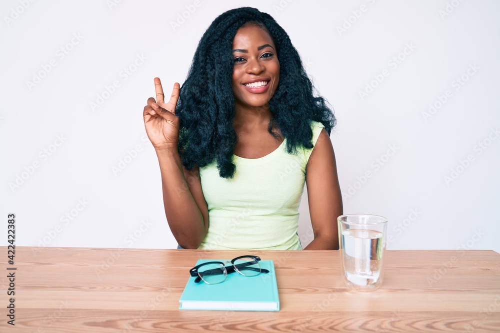 Beautiful african woman sitting on the table stuying for university smiling with happy face winking at the camera doing victory sign. number two. Beautiful african woman sitting on the table stuying for university smiling with happy face winking at the camera doing victory sign. number two.