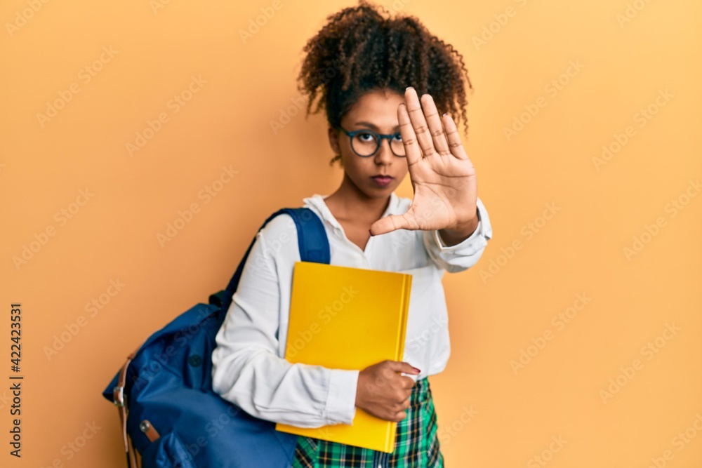 Beautiful african american woman with afro hair wearing school bag and holding books with open hand doing stop sign with serious and confident expression, defense gesture Beautiful african american woman with afro hair wearing school bag and holding books with open hand doing stop sign with serious and confident expression, defense gesture