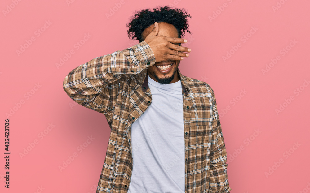 Young african american man with beard wearing casual clothes and glasses smiling and laughing with hand on face covering eyes for surprise. blind concept. Young african american man with beard wearing casual clothes and glasses smiling and laughing with hand on face covering eyes for surprise. blind concept.