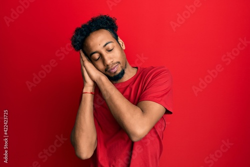 Young african american man with beard wearing casual red t shirt sleeping tired dreaming and posing with hands together while smiling with closed eyes. © Krakenimages.com