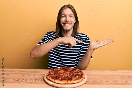 Handsome caucasian man with long hair eating tasty pepperoni pizza amazed and smiling to the camera while presenting with hand and pointing with finger.