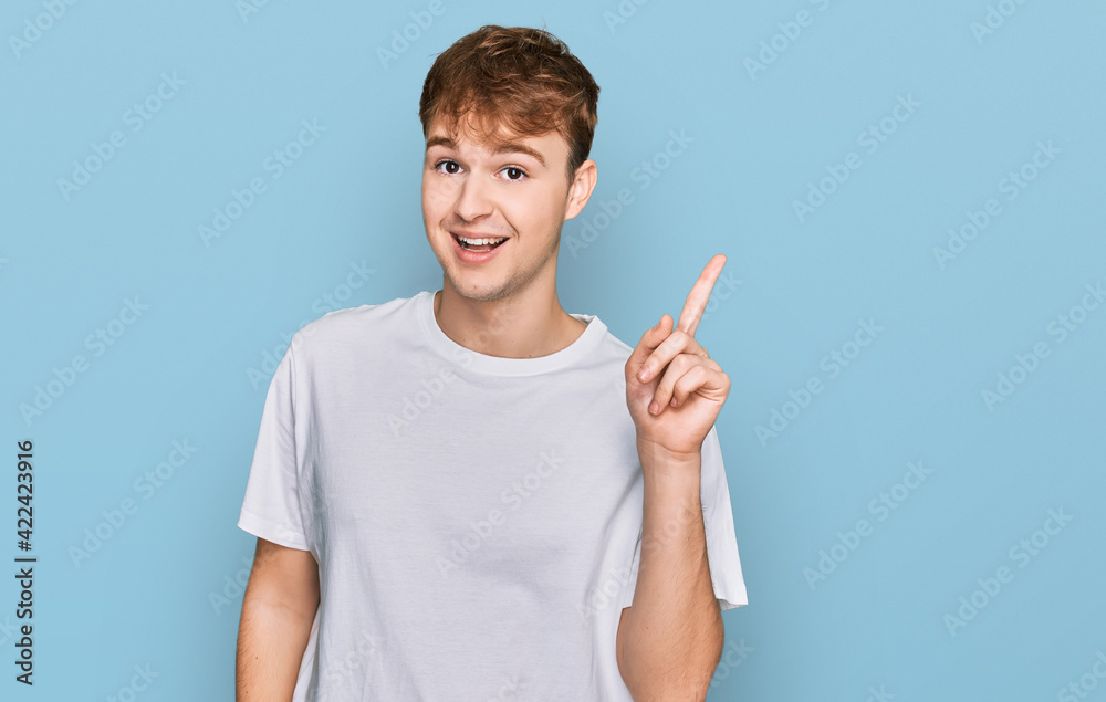 Young caucasian man wearing casual white t shirt showing and pointing up with finger number one while smiling confident and happy. Young caucasian man wearing casual white t shirt showing and pointing up with finger number one while smiling confident and happy.