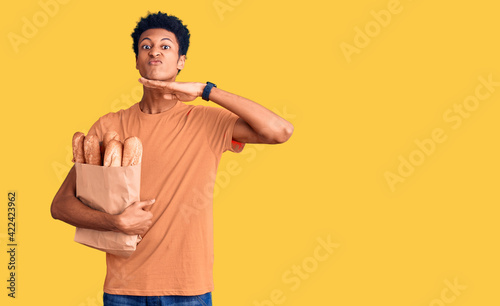 Young african american man holding paper bag with bread cutting throat with hand as knife, threaten aggression with furious violence