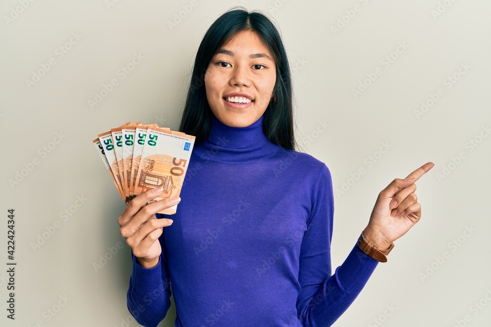 Young asian woman holding bunch of 50 euro banknotes smiling happy pointing with hand and finger to the side Young asian woman holding bunch of 50 euro banknotes smiling happy pointing with hand and finger to the side