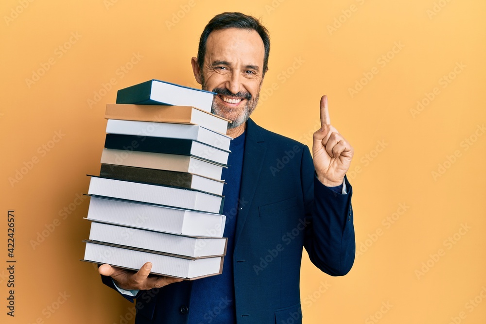 Middle age hispanic man holding a pile of books smiling with an idea or question pointing finger with happy face, number one Middle age hispanic man holding a pile of books smiling with an idea or question pointing finger with happy face, number one