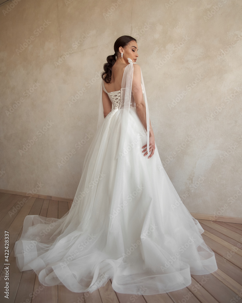Full length portrait of young beautiful woman wearing white wedding dress. Elegant bride standing and posing against beige wall Full length portrait of young beautiful woman wearing white wedding dress. Elegant bride standing and posing against beige wall