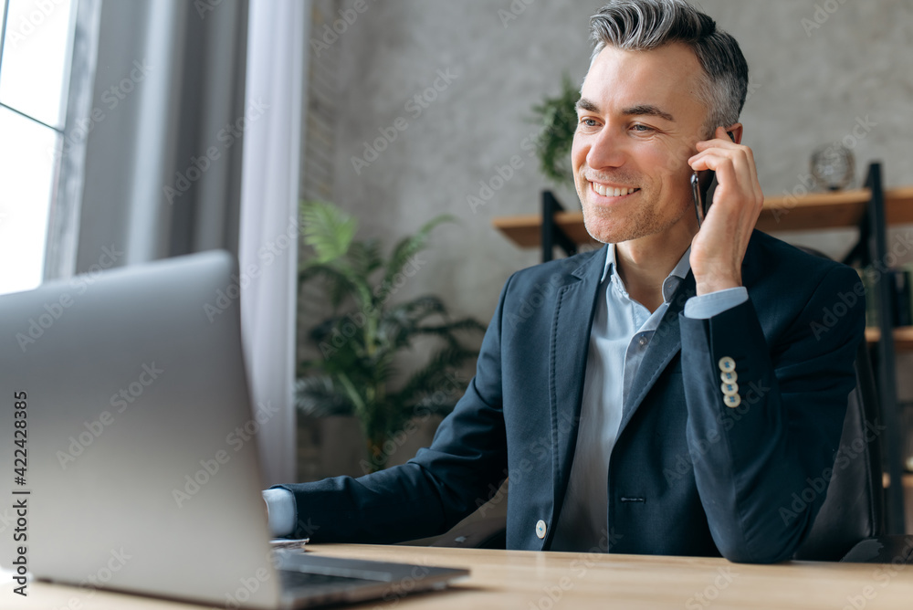 Busy attractive successful business leader sits at workplace in office, in a formal suit, works at a laptop, communicates with a client or employee by smartphone, smiles friendly looks at laptop