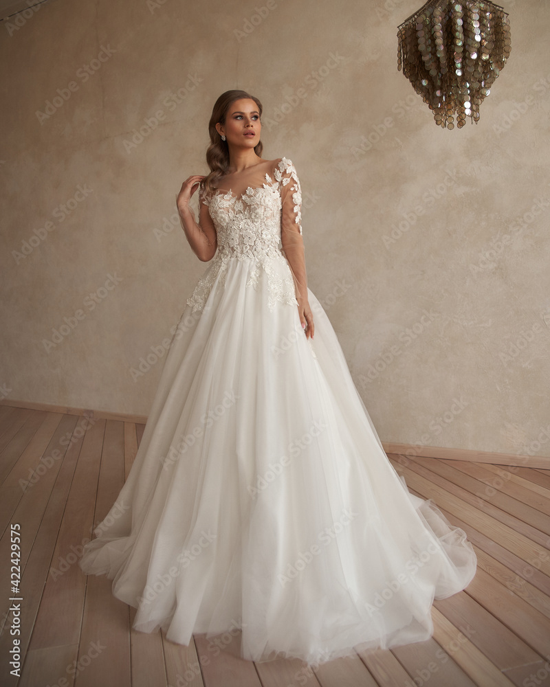 Fototapeta premium Full length portrait of young beautiful woman wearing white wedding dress. Elegant bride standing and posing against beige wall