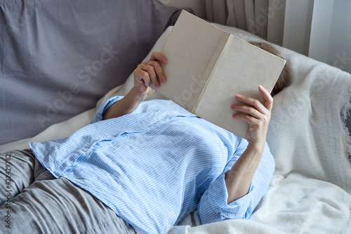 Senior woman in home clothes is reading a book while lying on her back on the couch, covering her face.