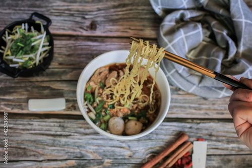 Hand uses chopsticks to pickup tasty egg noodle with pork and vegetable in five spices black soup on wooden table top view- Asian food style.