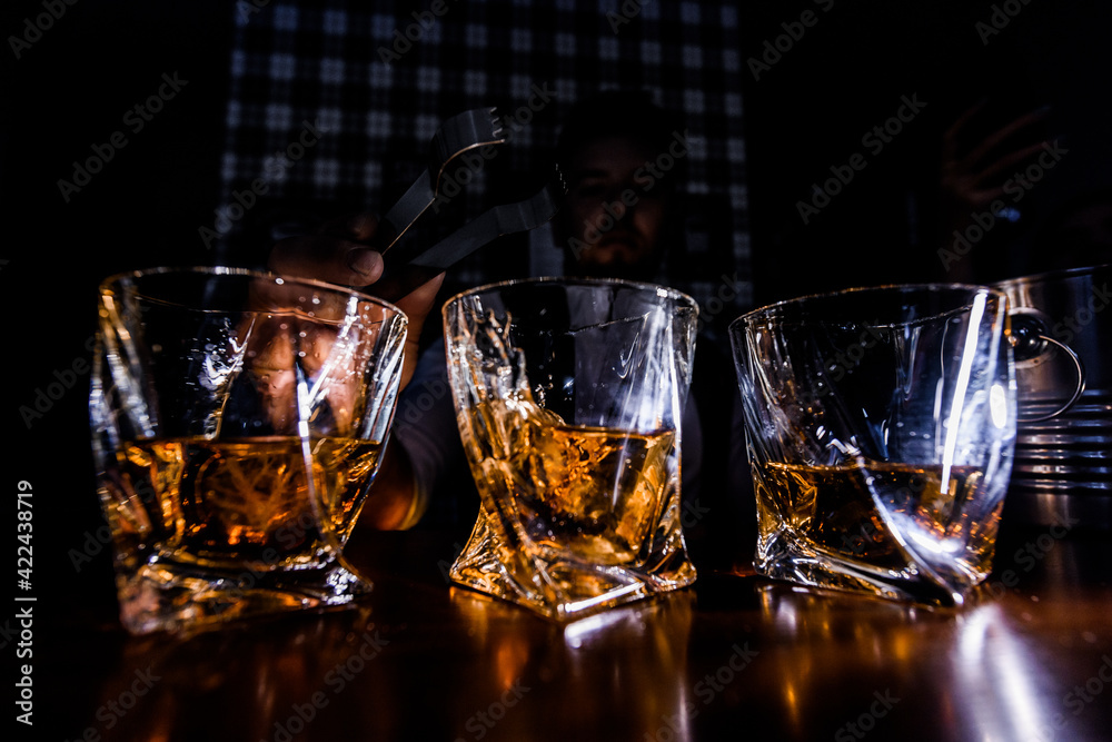 Front view of man puting ice cubes into glasses with whiskey on the dark light background. Front view of man puting ice cubes into glasses with whiskey on the dark light background.