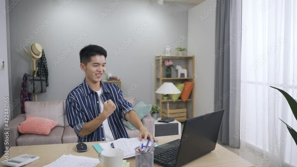 asian man sitting down at the table and looking at computer to celebrate good work results by punching fist into palm is feeling motivated in his job at home. asian man sitting down at the table and looking at computer to celebrate good work results by punching fist into palm is feeling motivated in his job at home.