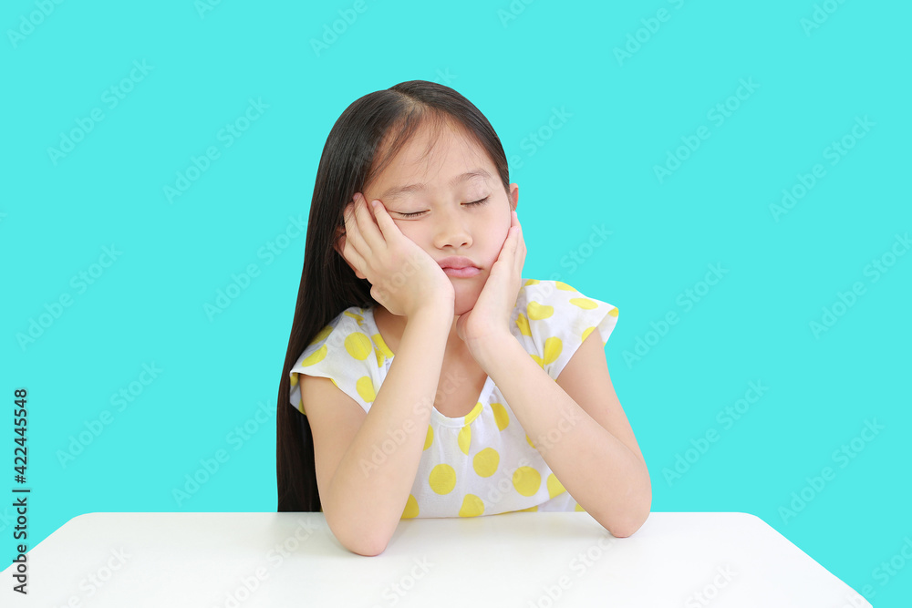 Sleepy little Asian child girl resting chin on hands on table against cyan background. Tired Schoolgirl at desk