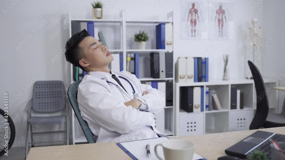 tired asian male medical worker is throwing back his head with closed eyes and folded arms while sitting resting in the office chair at a modern bright clinic. tired asian male medical worker is throwing back his head with closed eyes and folded arms while sitting resting in the office chair at a modern bright clinic.