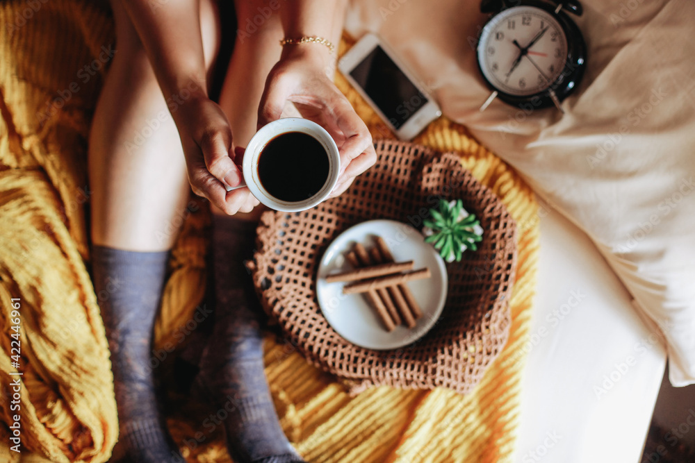 Top view of hand holding a cup of coffee with tasty snack for breakfast and clock showing 7 o'clock and feet in warm sock on the bed - obrazy, fototapety, plakaty 