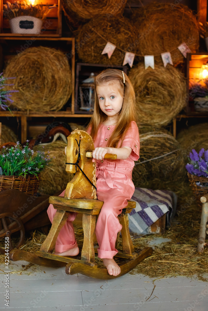 A pretty girl sits on a wooden rocking horse in the hay among baskets of lavender flowers. The blonde girl is dressed in a pink jumpsuit.