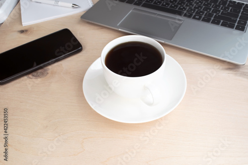 A close-up of a white coffee cup stands on a wooden table against a background of a black mobile phone and a gray laptop.