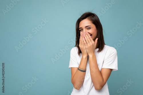Portrait of happy positive young beautiful brunette woman with sincere emotions wearing casual white t-shirt for mockup isolated on blue background with copy space and laughing covering mouth