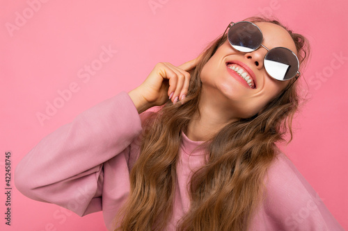Shot of beautiful positive smiling happy young dark blonde curly woman isolated over pink background wall wearing casual pink sport clothes and stylish sunglasses looking up