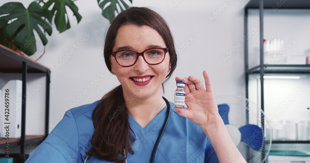 Beautiful young doctor woman in blue nurse uniform smiling at camera showing vaccine flask. Hope in fighting coronavirus Beautiful young doctor woman in blue nurse uniform smiling at camera showing vaccine flask. Hope in fighting coronavirus