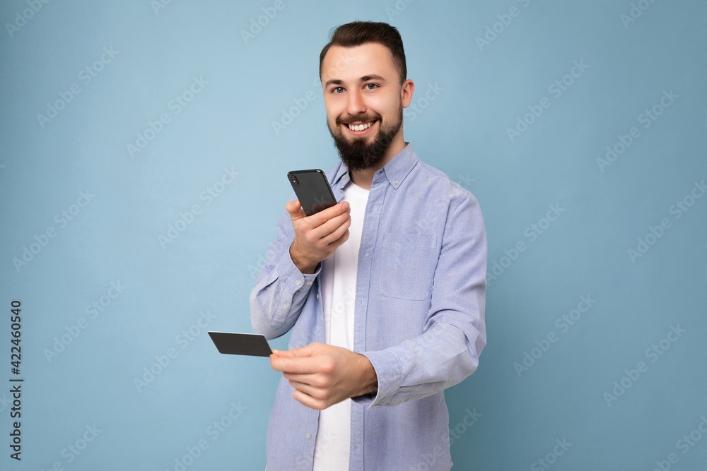 Photo of good looking attractive smiling brunet unshaven young man wearing casual blue shirt and white t-shirt isolated over blue background wall holding credit card and using mobile phone taking Photo of good looking attractive smiling brunet unshaven young man wearing casual blue shirt and white t-shirt isolated over blue background wall holding credit card and using mobile phone taking