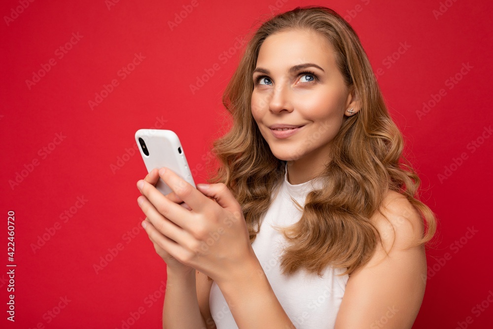 Closeup portrait of happy pretty young blonde woman wearing white t-shirt isolated over red background using smartphone and chatting via mobile phone looking to the side and thinking Closeup portrait of happy pretty young blonde woman wearing white t-shirt isolated over red background using smartphone and chatting via mobile phone looking to the side and thinking