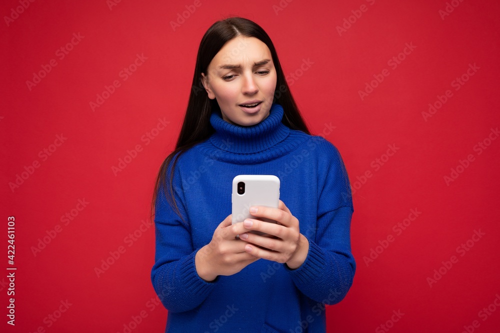 Thoughtful attractive good looking young brunette woman wearing stylish blue warm sweater poising isolated on red background with empty space holding in hand and using mobile phone reading news online Thoughtful attractive good looking young brunette woman wearing stylish blue warm sweater poising isolated on red background with empty space holding in hand and using mobile phone reading news online