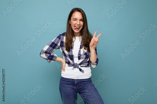 Young positive happy beautiful winsom brunette woman with sincere emotions wearing check shirt poising isolated over blue background with copy space and showing peace gesture