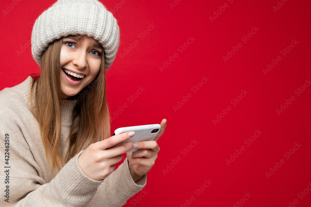 Closeup portrait of Beautiful emotionaloverjoyed young blonde woman wearing casual beige sweater and beige hat isolated over red background holding in hand and using mobile phone playing online games Closeup portrait of Beautiful emotionaloverjoyed young blonde woman wearing casual beige sweater and beige hat isolated over red background holding in hand and using mobile phone playing online games