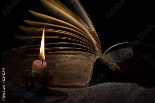 A candle burns in the dark. In the background there is an old book with open pages. The concept of mysticism and secret knowledge, esotericism. Selective focus on a burning candle. photo