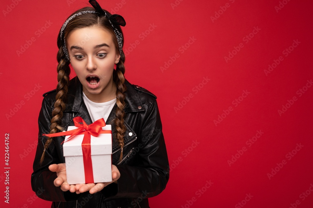 Photo of beautiful happy amazed brunette girl isolated over red background wall wearing white casual t-shirt and stylish black leather jacket holding white gift box with red ribbon and looking at Photo of beautiful happy amazed brunette girl isolated over red background wall wearing white casual t-shirt and stylish black leather jacket holding white gift box with red ribbon and looking at
