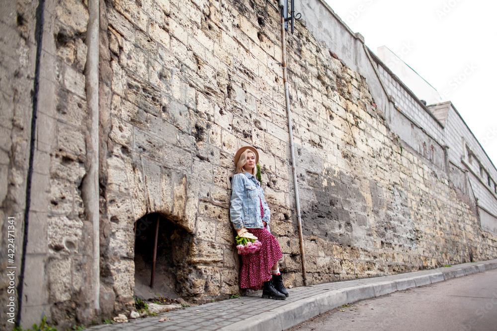 Fashion style woman dressed jeans jacket, hat and red dress walking in the street of old city with bouquet of flowers