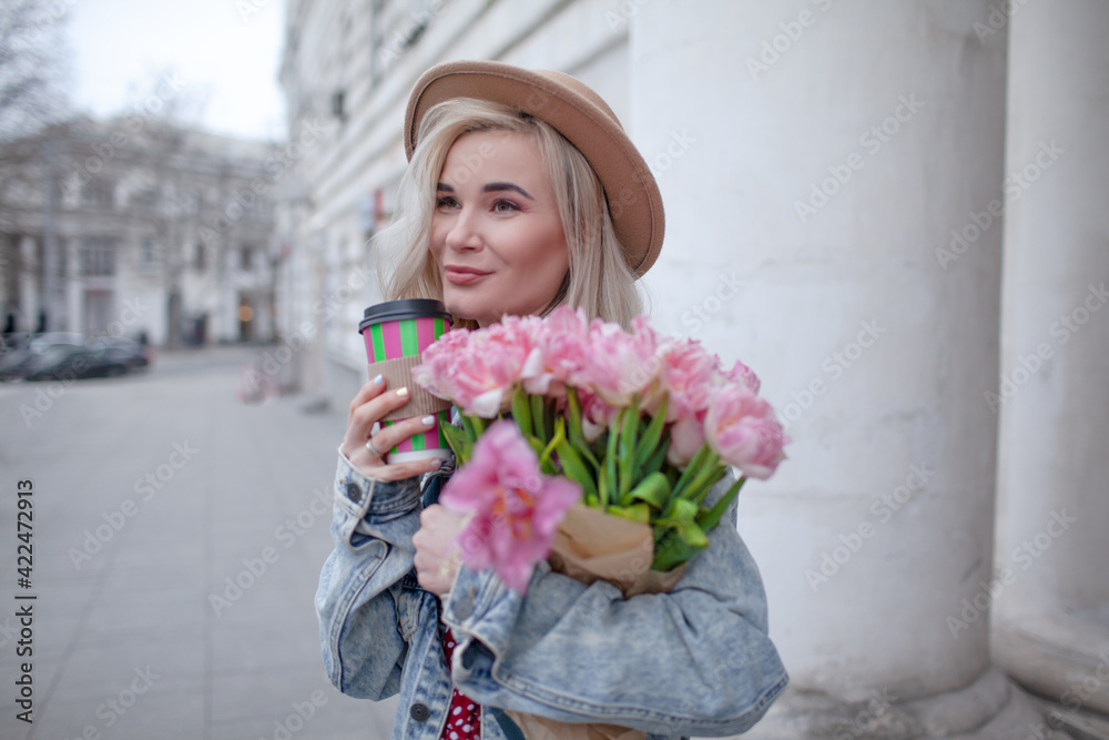 Fototapeta premium Beautiful woman in fashion clothes walking in the street with bouquete of flowers and coffee or tea to go
