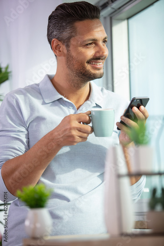 cheerful man stood by window holding coffee and smartphone