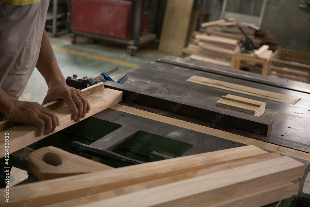 A young male carpenter builder in a gray T-shirt and working overall equals a wooden bar with a milling machine in the workshop, in the background wooden boards, in the workshop