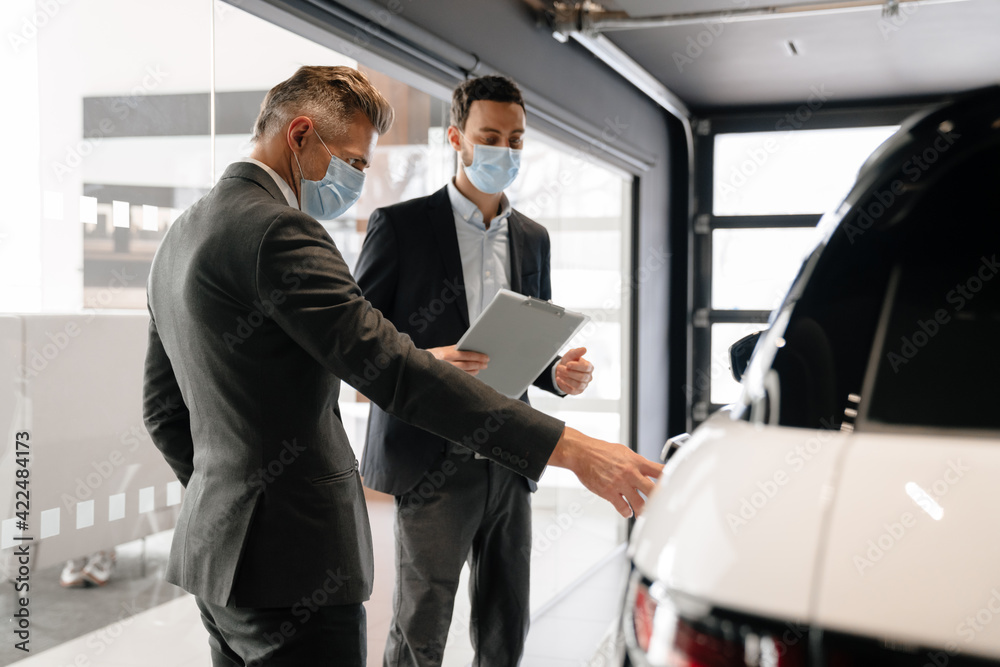 Mid white manager in face mask showing car to customer in showroom Mid white manager in face mask showing car to customer in showroom