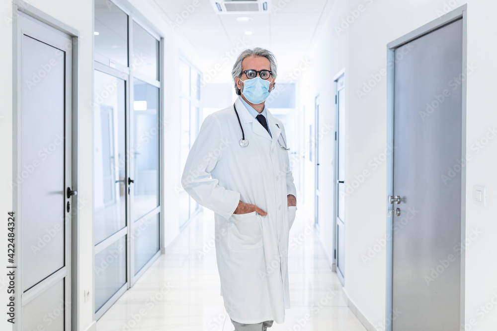 Confident doctor wearing protective face mask while standing in a hallway at the hospital. Senior doctor, surgeon, medic staff professional portrait.