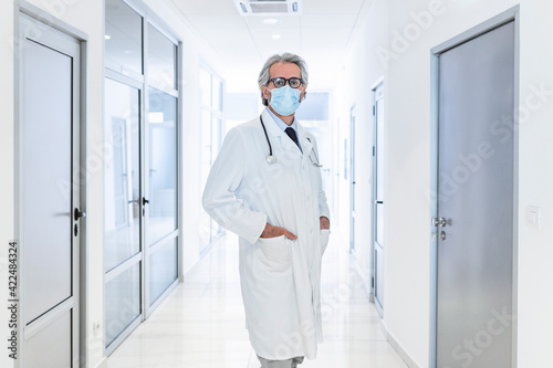Confident doctor wearing protective face mask while standing in a hallway at the hospital. Senior doctor, surgeon, medic staff professional portrait.