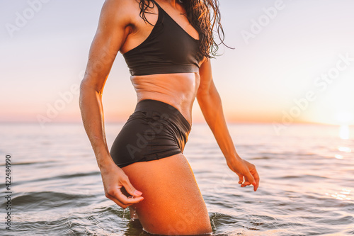 Anonymous sporty slim body of woman posing in calm sea at sunset in black sportswear. Liposuction, diet, abs