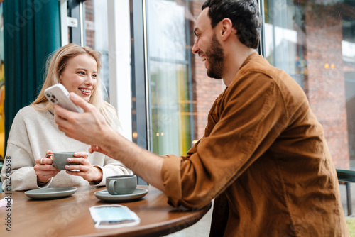 Smiling man and woman talking and using cellphone while drinking coffee