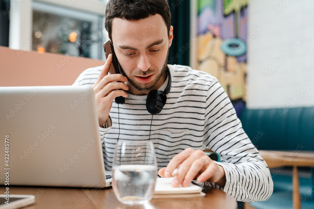 Focused young man talking on cellphone while working with laptop in cafe Focused young man talking on cellphone while working with laptop in cafe
